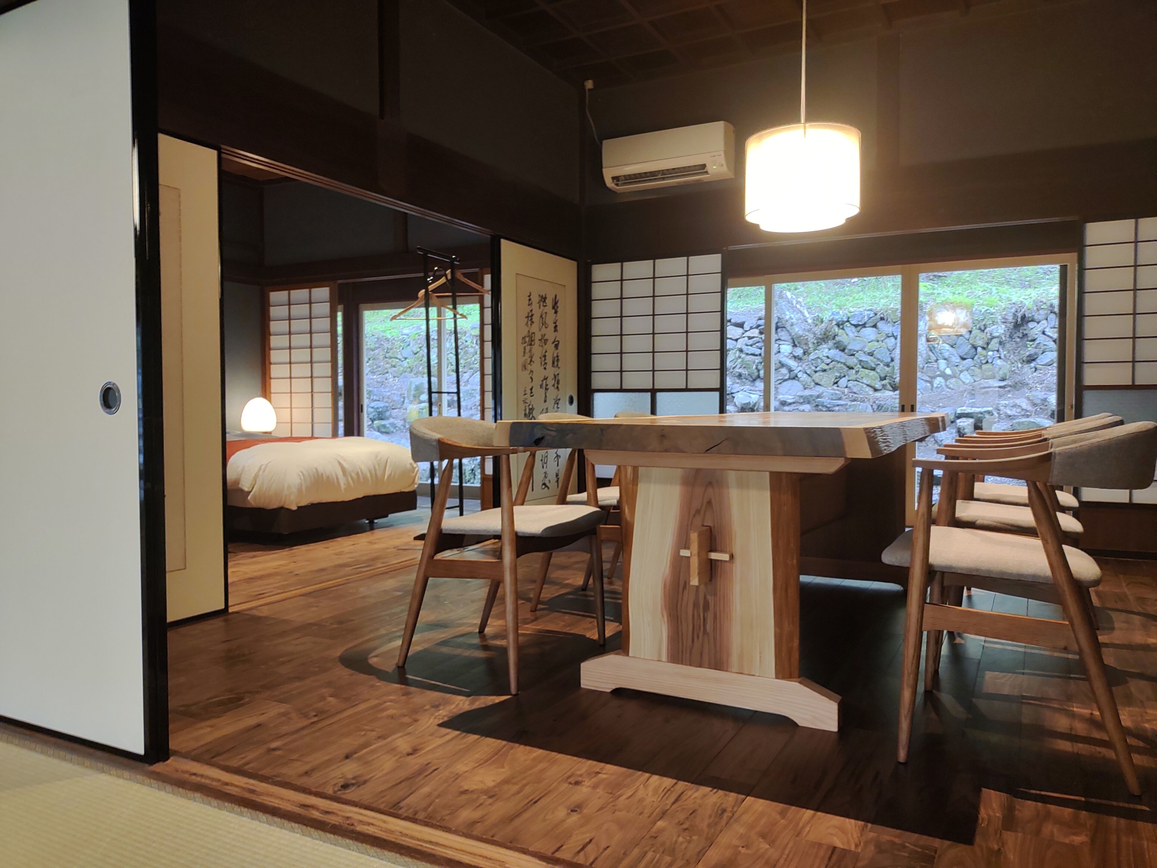 A traditional Japanese-style room with tatami flooring, a low wooden table with chairs, a bed with a white bedspread, and a view of a garden through large windows.