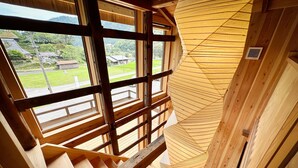 Een houten interieur met een trap naar een vide, een raam met uitzicht op een groen landschap, en een tafeltje met kopjes en een theepot.