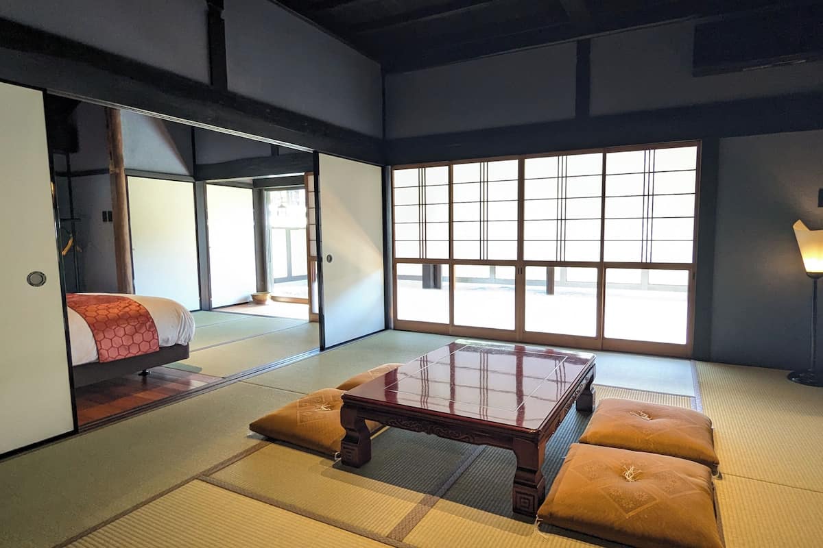 Une pièce de style japonais traditionnel, avec un sol recouvert de tatamis, une basse table en bois, des coussins, des portes coulissantes et une vue dégagée sur l’extérieur grâce à de grandes fenêtres.