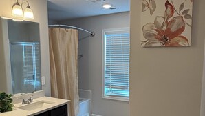 A bathroom with a double sink vanity, a shower with a beige curtain, a large mirror, and a window with blinds.