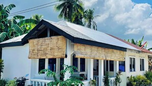 A single-story house with a corrugated metal roof, white walls, and a porch with a bamboo awning. There are several potted plants around the house, and a motorcycle parked in front. - Islescape Siargao (General Luna)