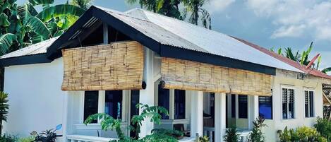 A single-story house with a corrugated metal roof, white walls, and a porch with a bamboo awning. There are several potted plants around the house, and a motorcycle parked in front.