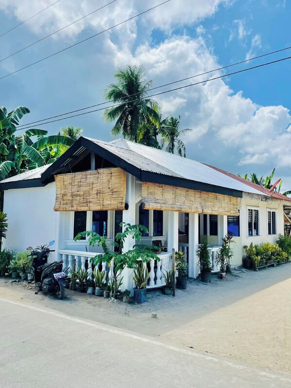 A single-story house with a corrugated metal roof, white walls, and a porch with a bamboo awning. There are several potted plants around the house, and a motorcycle parked in front.