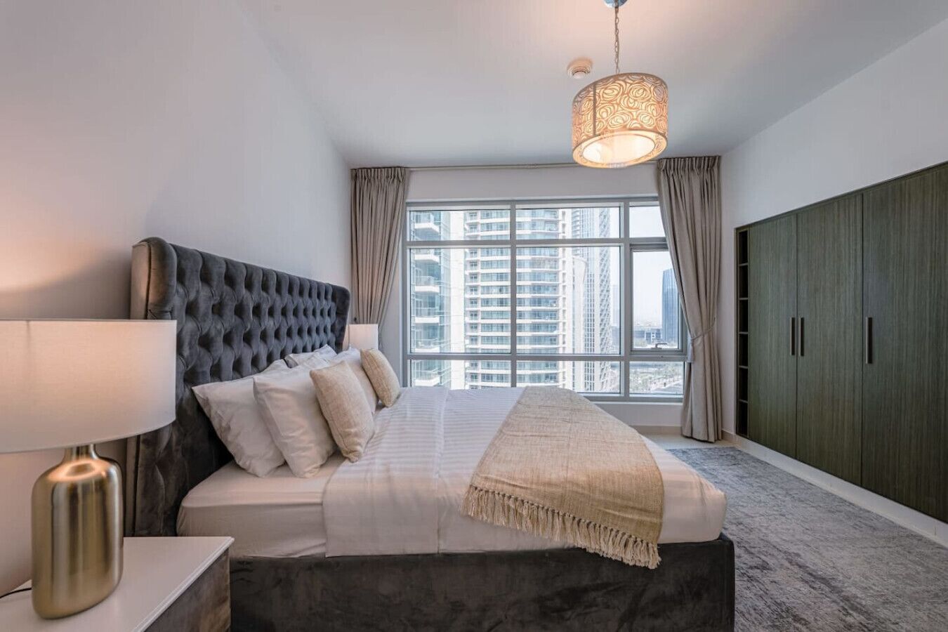 Ein modernes Schlafzimmer mit einer gepolsterten Kopfteil, einem großen Bett mit beiger Tagesdecke, einer Hängeleuchte und Blick auf hohe Gebäude durch das Fenster.