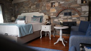 A bedroom with a stone wall, a bed with a blue bedspread, a bedside table with a lamp, and a sofa. - Domatia Stone Hotel (Soke)