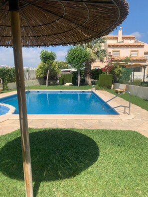 Una piscina con sombrilla de paja, rodeada de césped cuidado y una valla.