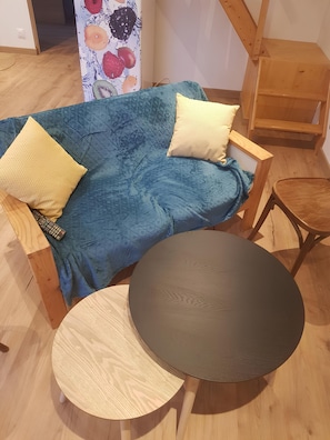 Ein Zimmer mit einem blauen Sofa, zwei Kissen, einem runden Holztisch und einem Holzstuhl.