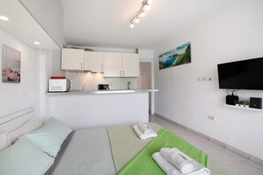 Pieni hotellihuone, jossa on sänky, vihreä pyyhe, valkoinen tyyny, pieni keittokomero, litteänäyttöinen televisio ja taulu seinällä.