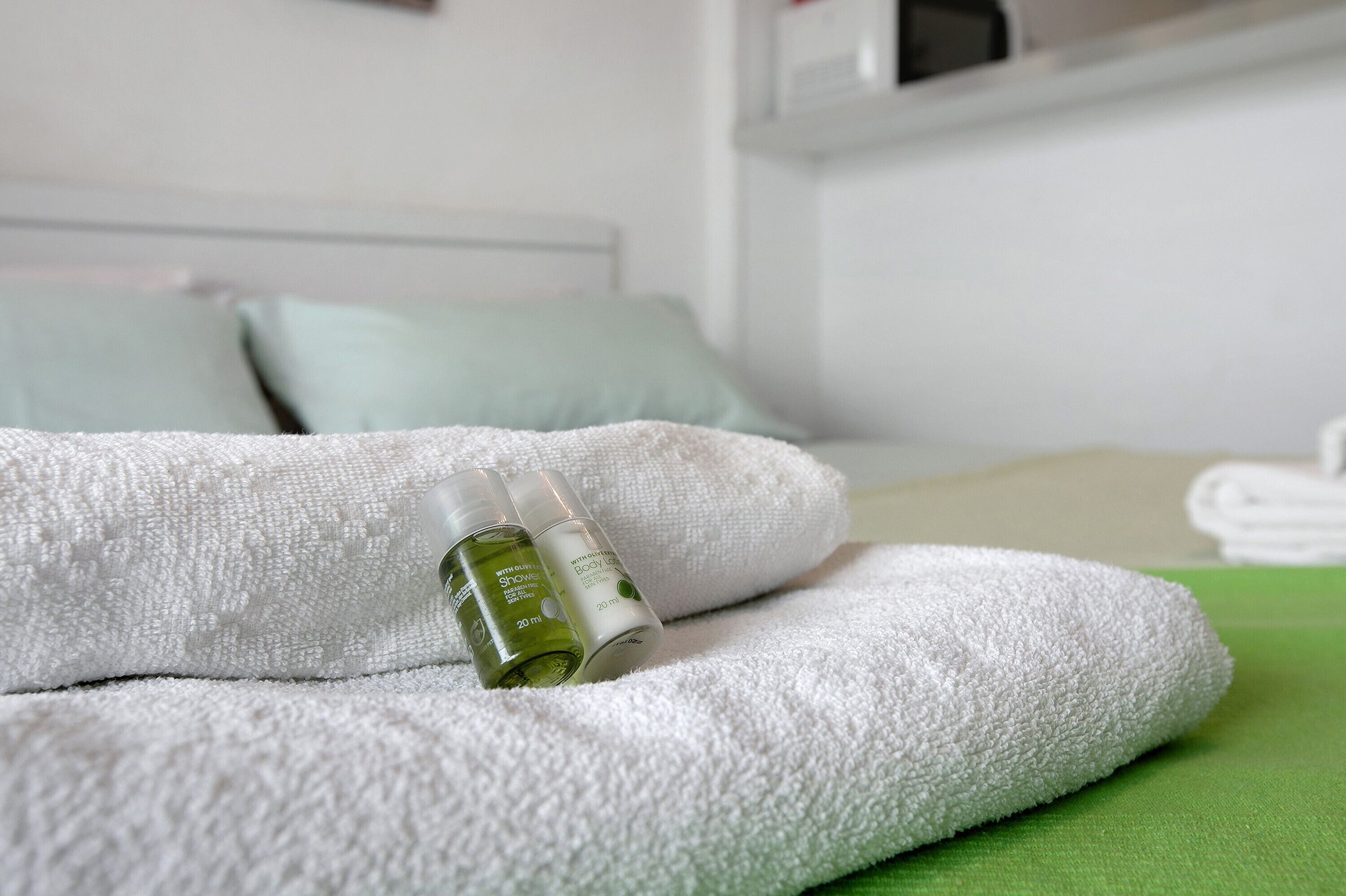 A neatly made bed with white and green linens, a bottle of body lotion, and a pillow.