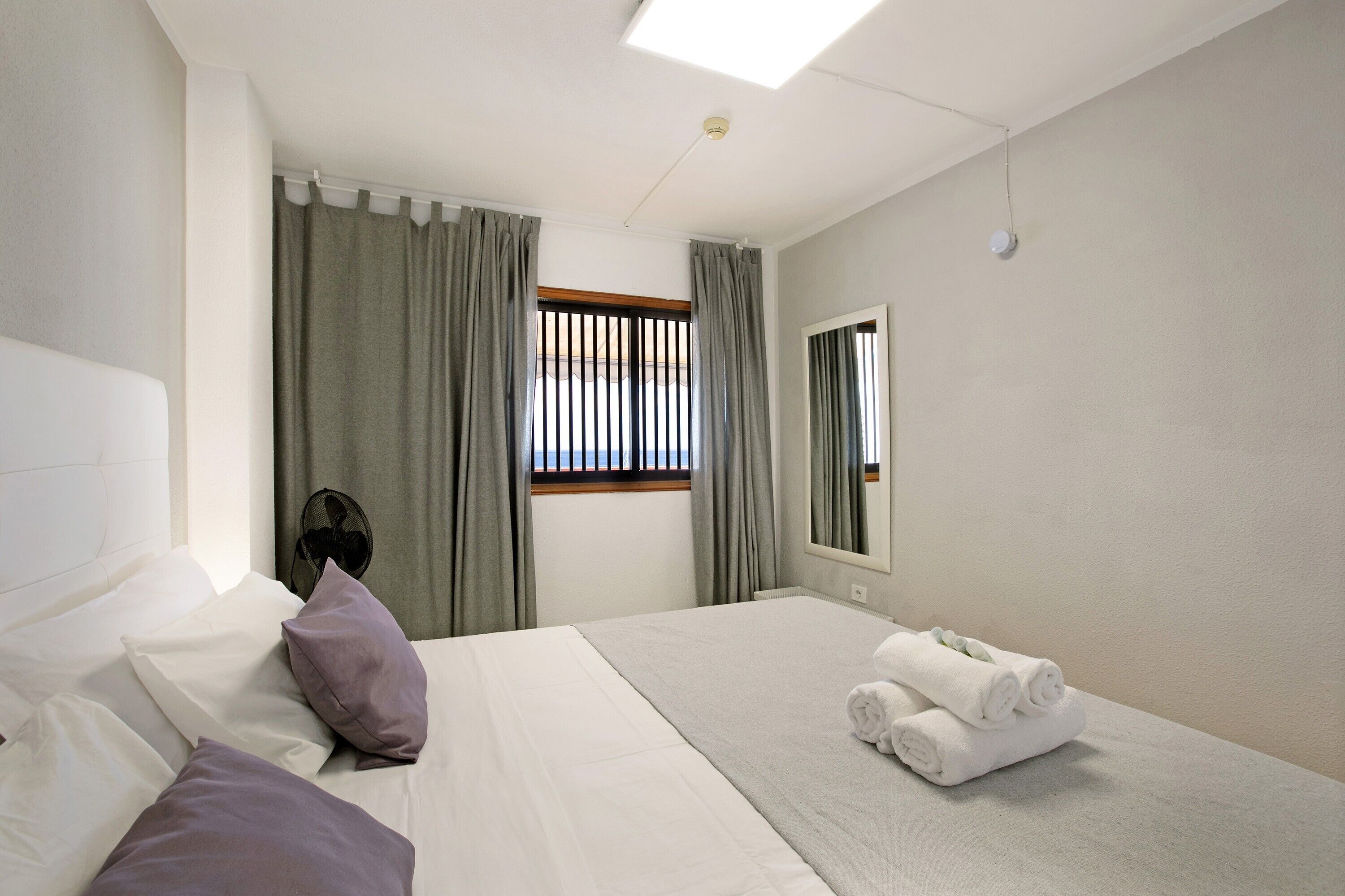 A hotel room with a bed, a window with curtains, a mirror, and a pair of folded towels on the floor.
