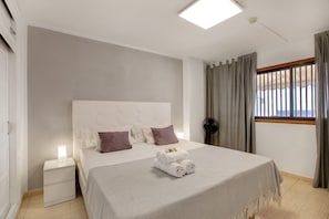 A modern hotel room with a large bed, a bedside table, a nightstand, a window with curtains, and a ceiling light. - Vacation Apartment 'Las Fuentes' with Sea View, Private Terrace and Wi-Fi (Arona)