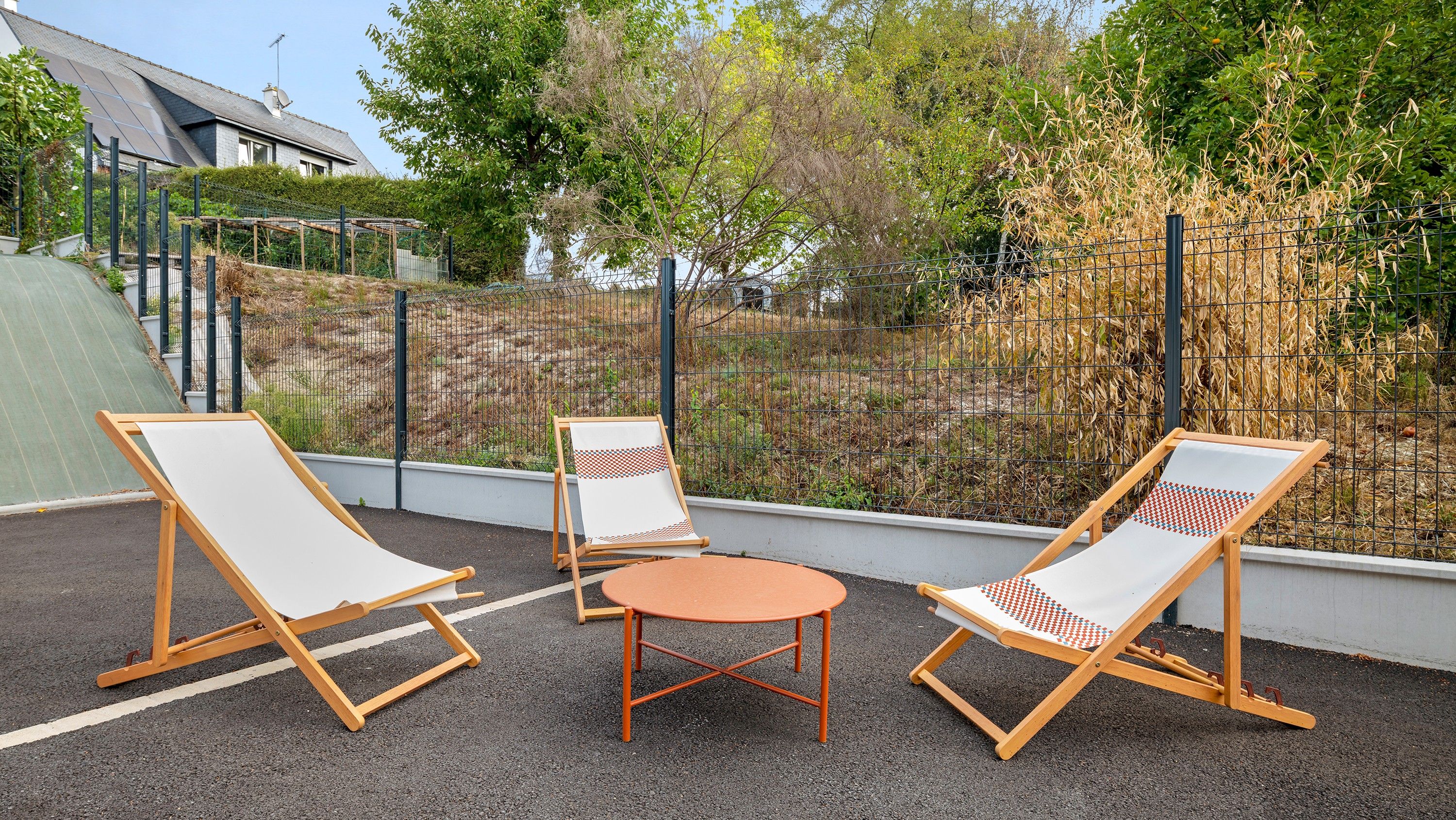 An outdoor seating area with two lounge chairs and a small table, surrounded by a metal fence and vegetation.