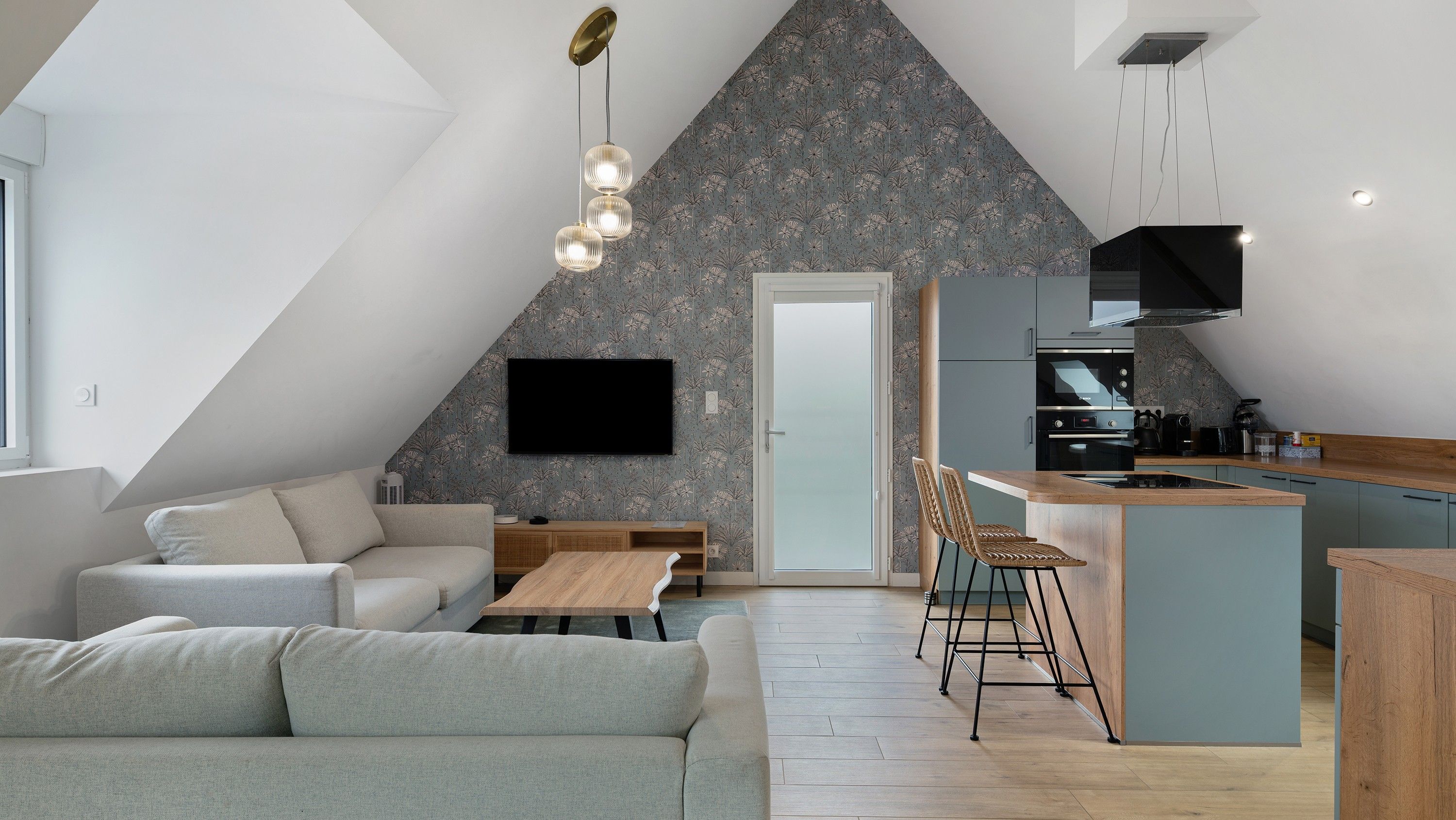Ein modernes Loft mit schrägem Dach, einem Essbereich mit Holztisch und Stühlen, einer Küche mit integrierter Kochinsel und einem an der Wand befestigten Flachbildfernseher.