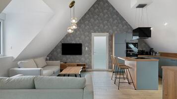 Ein modernes Loft mit schrägem Dach, einem Essbereich mit Holztisch und Stühlen, einer Küche mit integrierter Kochinsel und einem an der Wand befestigten Flachbildfernseher.