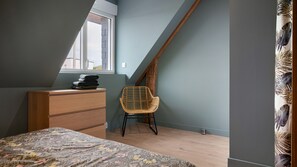 A bedroom with a sloped ceiling, a wooden dresser, a wicker chair, and a bed with a floral bedspread. - Apartment 'Appartement Centre - La Gacilly' with Wi-Fi (La Gacilly)