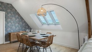 Un comedor con una mesa dispuesta para cuatro, una silla de ratán, una lámpara colgante de fibras naturales y vistas a través de un tragaluz.