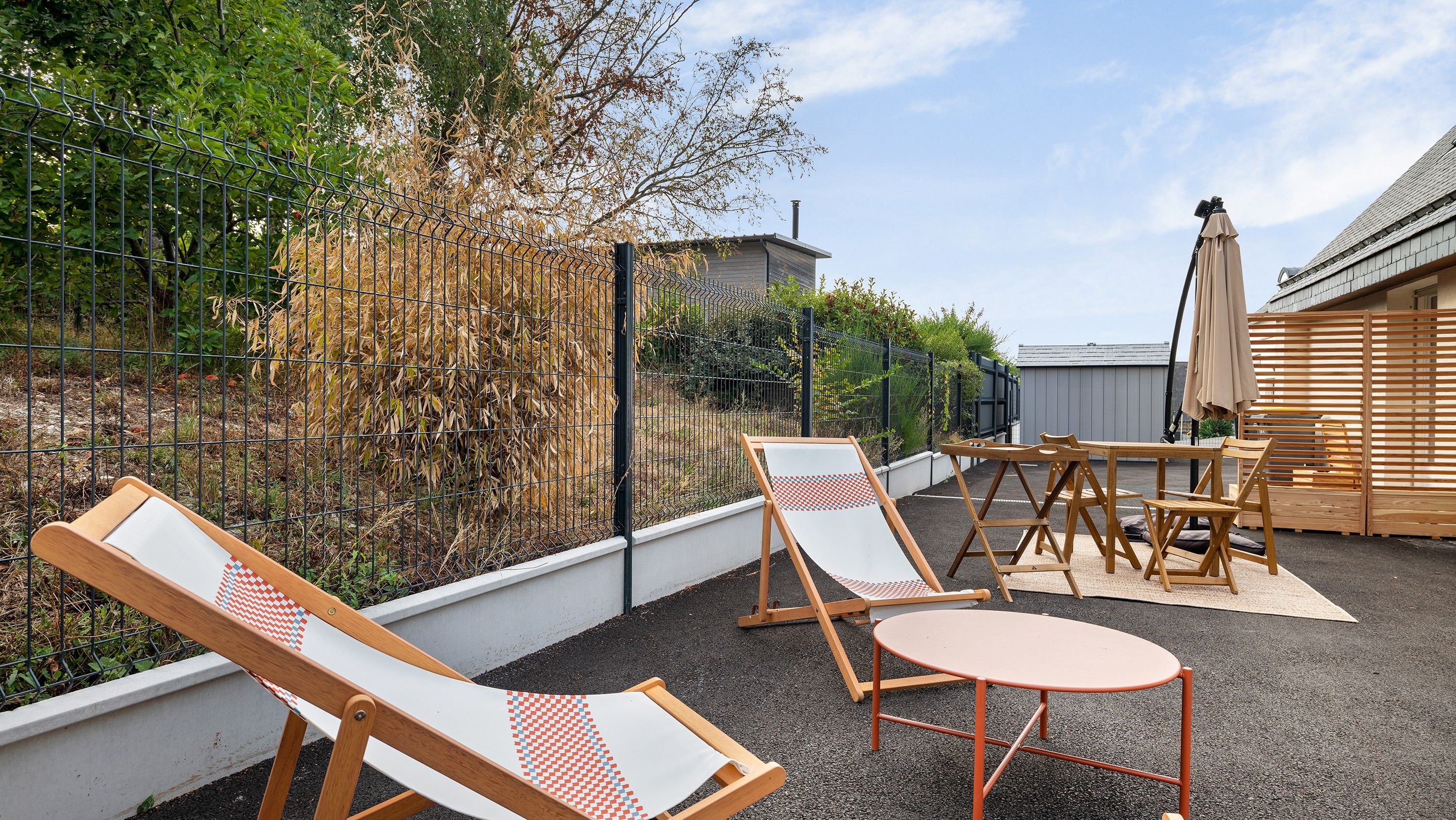 Eine Terrasse mit Gartenmöbeln, darunter zwei Liegestühle und ein Tisch, umgeben von einer Hecke und Bepflanzung.
