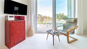 Une pièce avec un téléviseur, un meuble rouge portant l’inscription « POSTE », un vase en osier, une chaise grise en fibres tressées et une vue sur l’extérieur, visible à travers une porte-fenêtre coulissante.