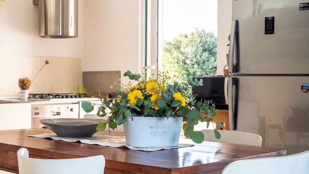 Une cuisine avec une table en bois, des chaises blanches, un réfrigérateur en acier inoxydable et une fenêtre donnant sur des arbres.