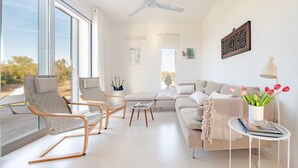 Un salon moderne comprenant un canapé, deux fauteuils, une table basse, un ventilateur de plafond et une vue dégagée sur l’extérieur grâce à de grandes fenêtres.