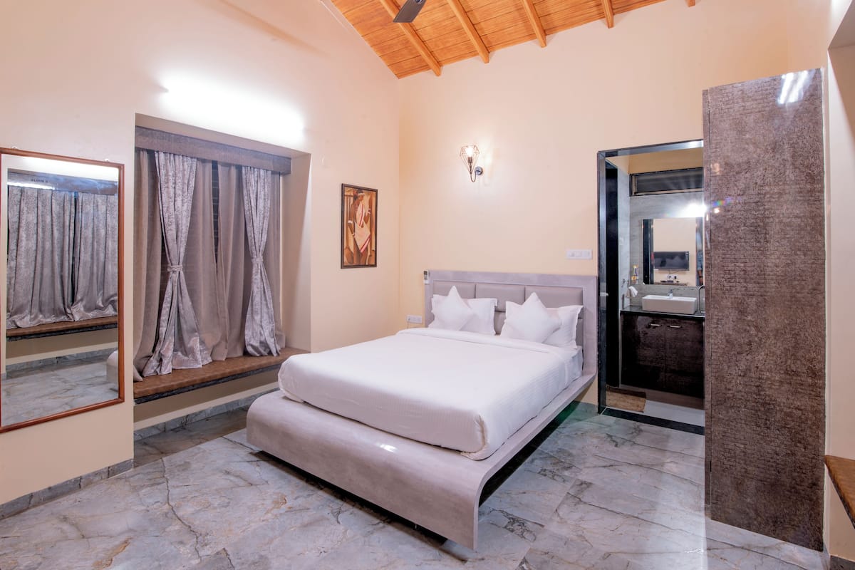 A bedroom with a bed, a large mirror, a window with curtains, and a bathroom with a sink and mirror.