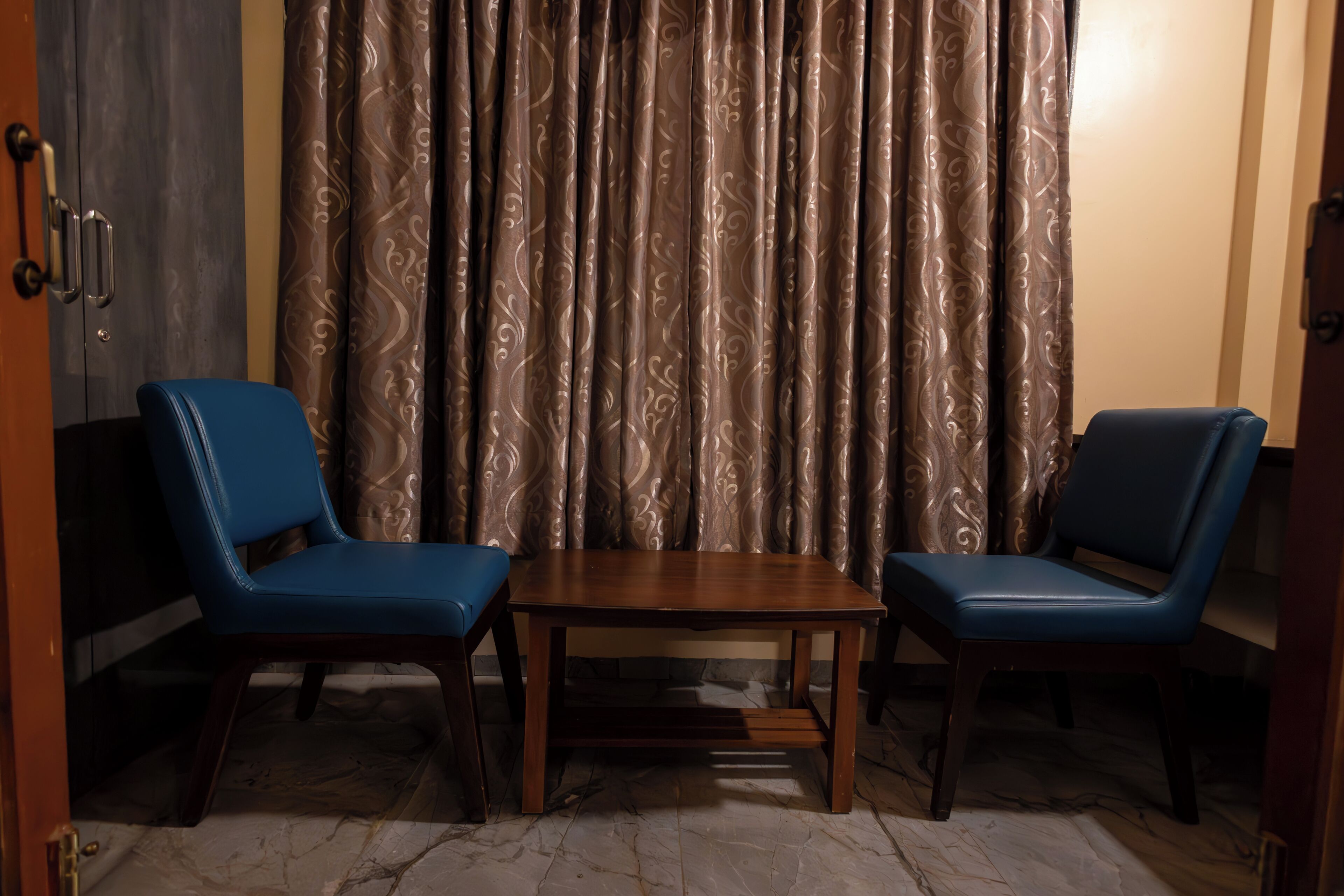 A room with two blue chairs, a wooden table, and a patterned curtain.