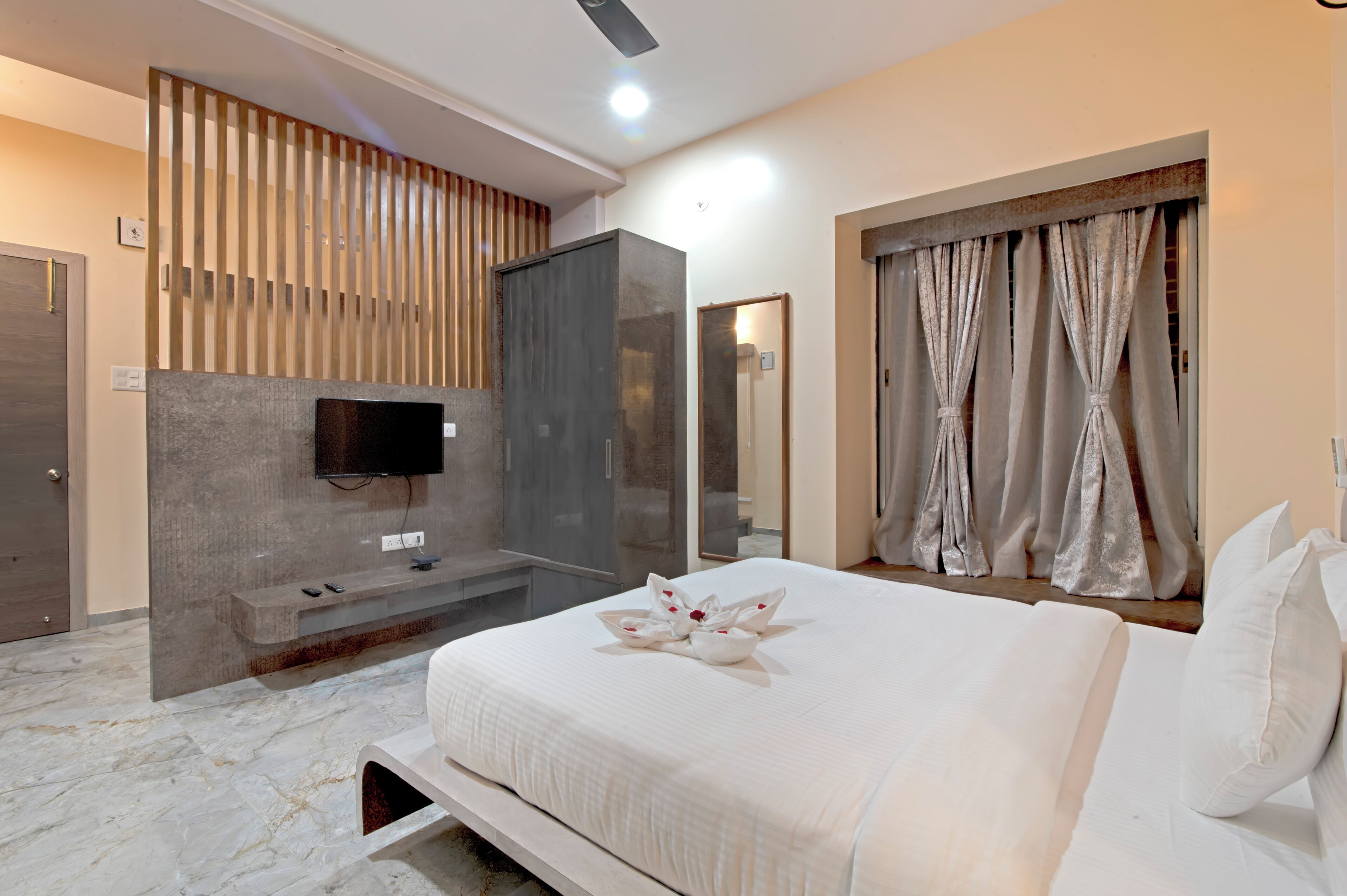 A modern hotel room with a large bed, a TV mounted on the wall, a seating area with a bench, and a shower area.