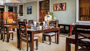 A dining area with wooden tables and chairs, a person serving, and a painting on the wall. - Astorian Grand Hotel (Naivasha)