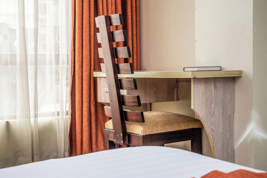 A hotel room with a bed, a wooden desk with a chair, and a window with curtains.