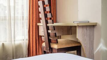 A hotel room with a bed, a wooden desk with a chair, and a window with curtains.