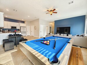 A modern living room with a pool table, a ceiling fan, and a TV. - Modern 6 Bedroom House with Tons of Games, BBQ, 2 85" TVs and Surround Sound! (Houston)