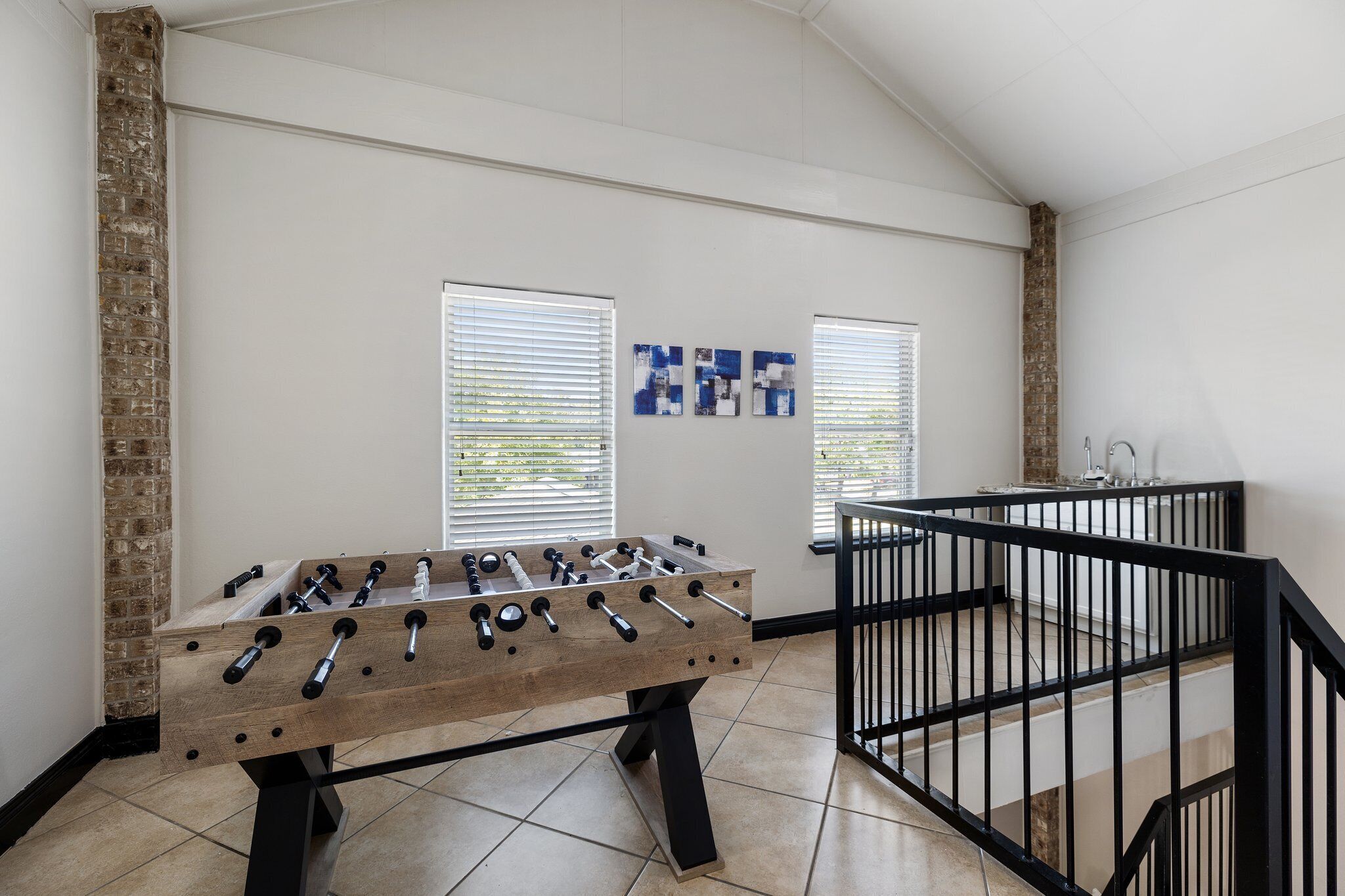 A room with a foosball table, a brick wall, and a window with blinds.