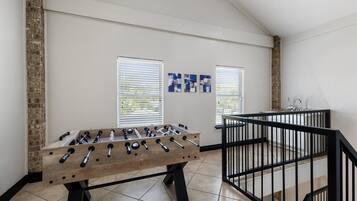 A room with a foosball table, a brick wall, and a window with blinds.
