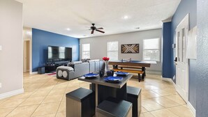 A spacious living room with a large TV, a dining area with a pool table, and a ceiling fan. - Modern 6 Bedroom House with Tons of Games, BBQ, 2 85" TVs and Surround Sound! (Houston)