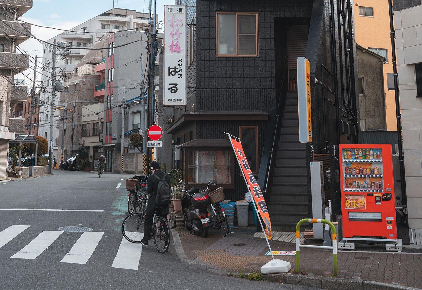 A street scene with a crosswalk, a no entry sign, a vending machine, and a building with signage.