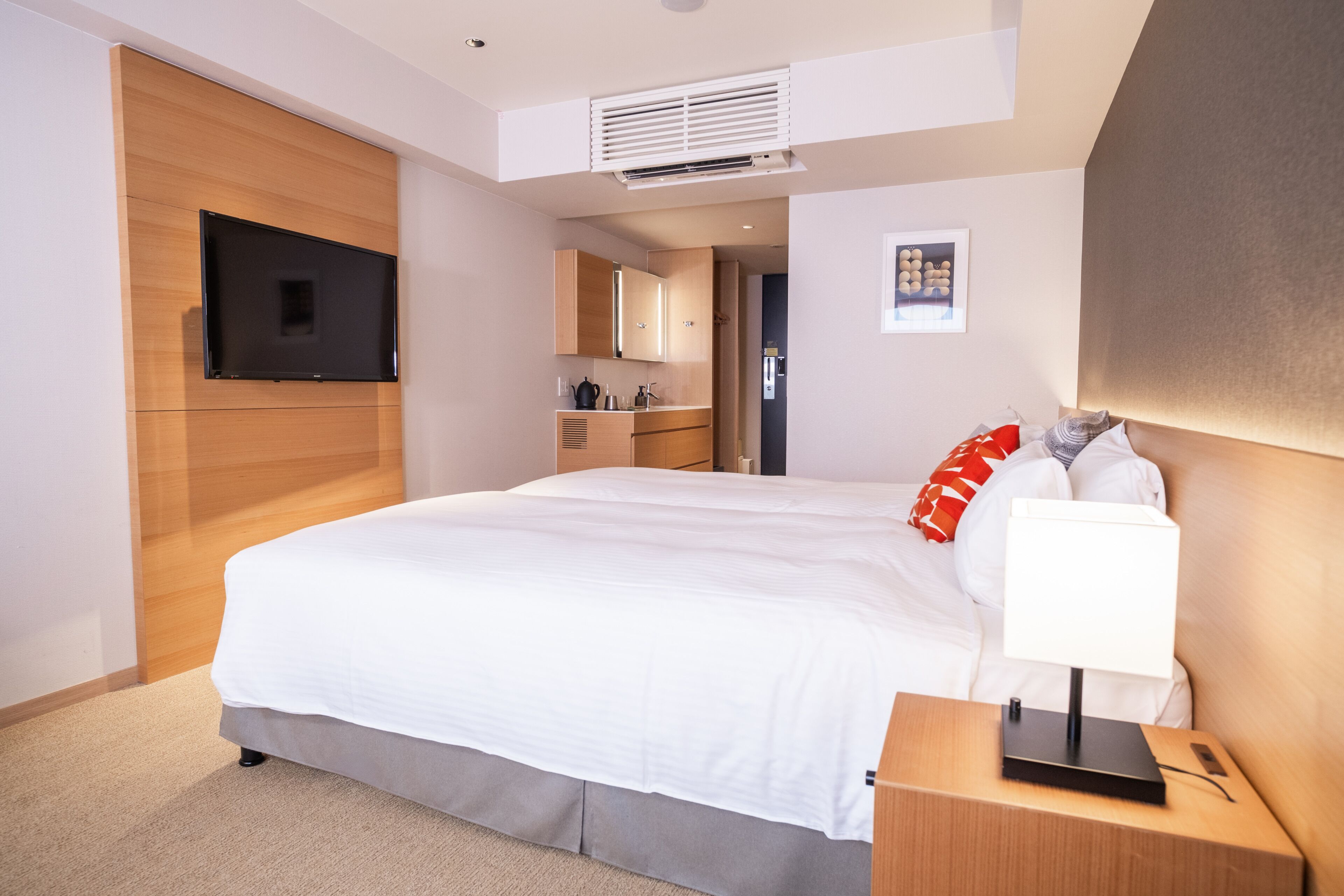 Quarto de hotel moderno com uma cama grande, mesa de cabeceira com abajur, televisão de tela plana na parede, cozinha compacta e um pequeno quadro na parede.