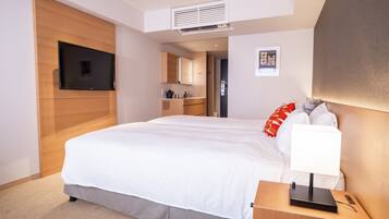 Quarto de hotel moderno com uma cama grande, mesa de cabeceira com abajur, televisão de tela plana na parede, cozinha compacta e um pequeno quadro na parede.