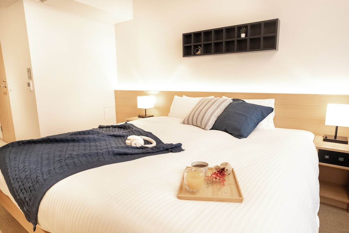 A neatly made bed with a white comforter, a dark blue blanket, and a tray with a glass of liquid and a small plant.