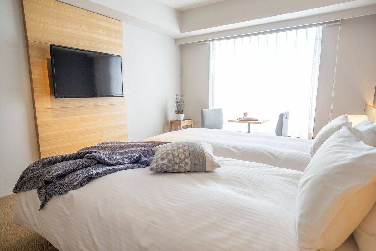 A hotel room with a bed, a flat-screen TV mounted on the wall, a small table with a lamp, and a window with curtains.