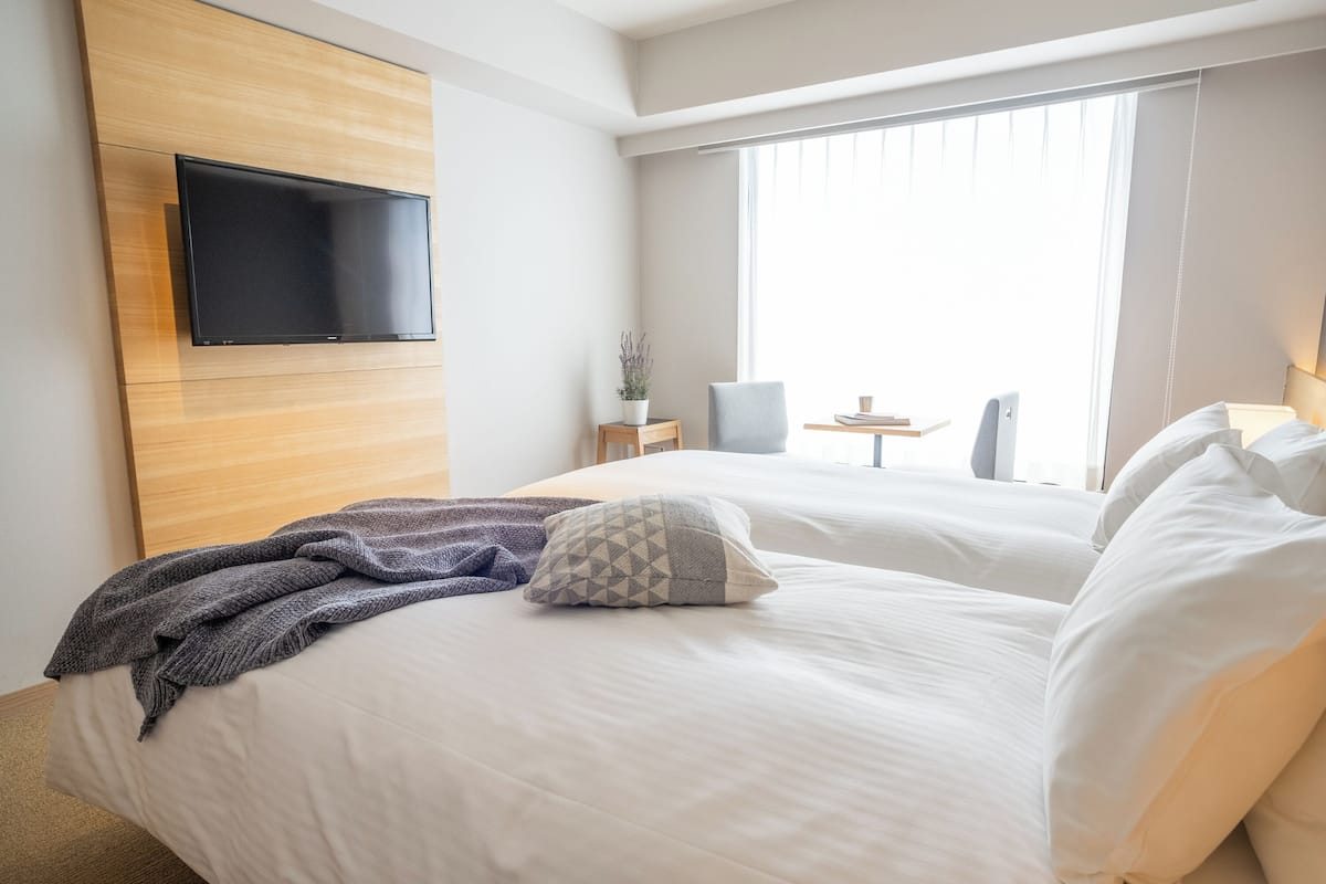 Ein Hotelzimmer mit Bett, flachbildfernseher an der Wand, einem kleinen Tisch mit Lampe und einem Fenster mit Vorhängen.