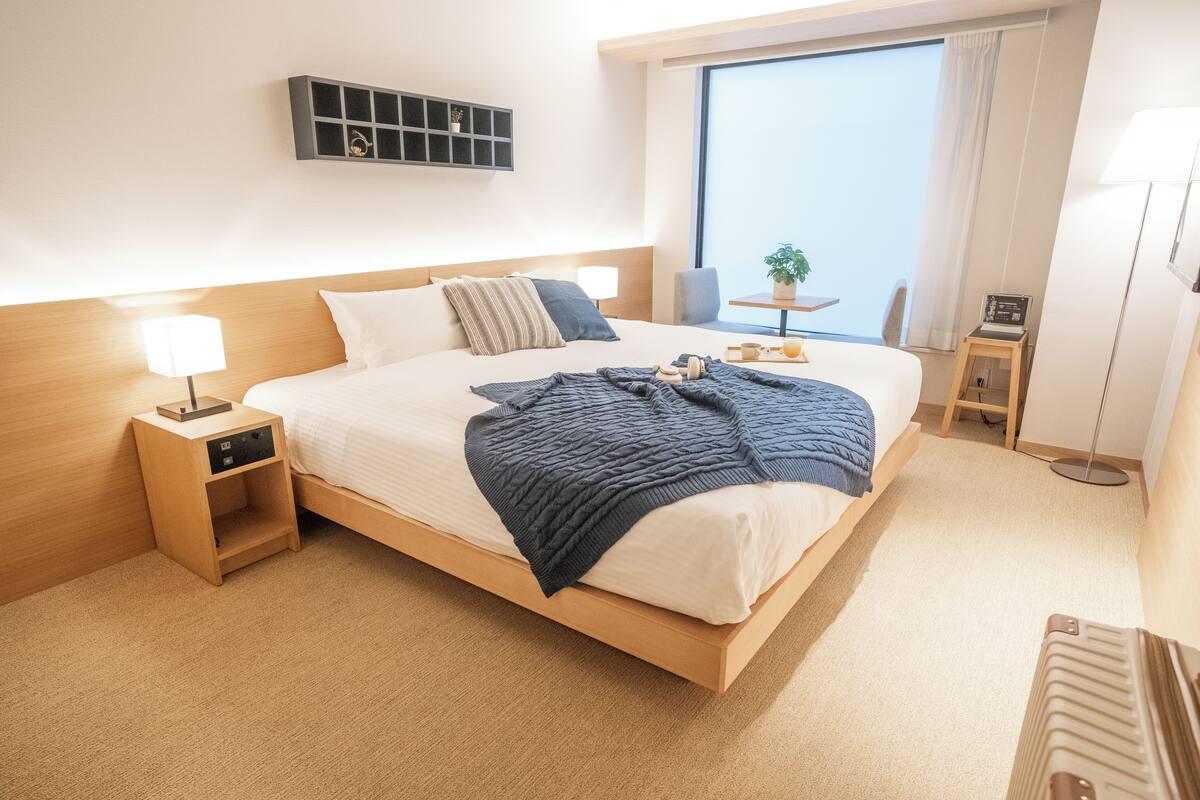 A modern hotel room with a large bed, bedside table with a lamp, a small table with a plant, and a view of the city through a large window.