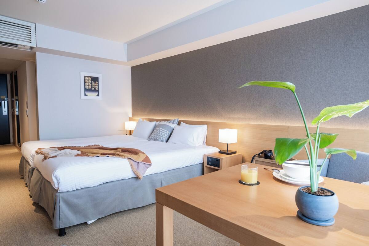 A hotel room with a bed, bedside table, lamp, and a potted plant.