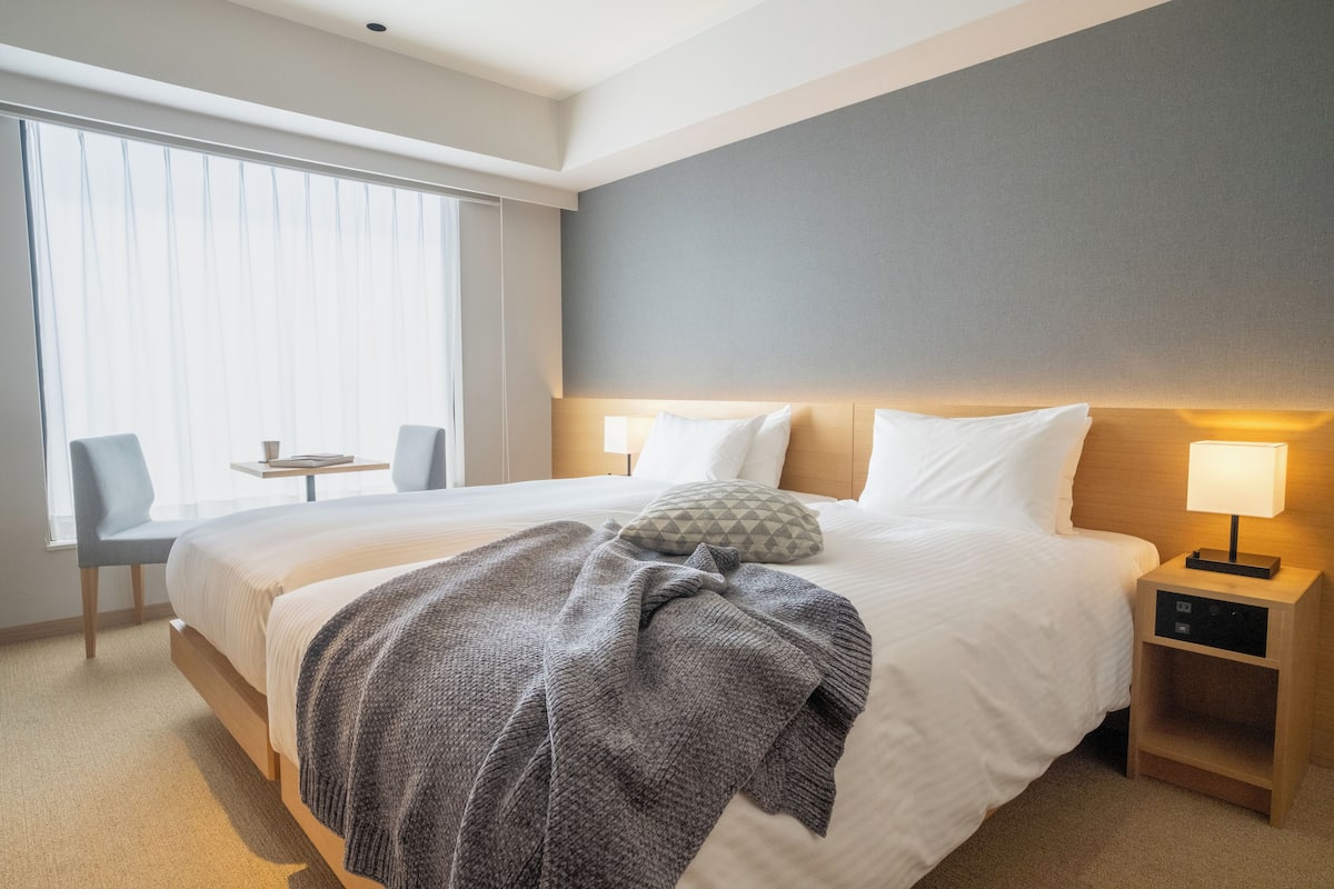 A hotel room with a large bed, a bedside table, a chair, and a window with blinds.