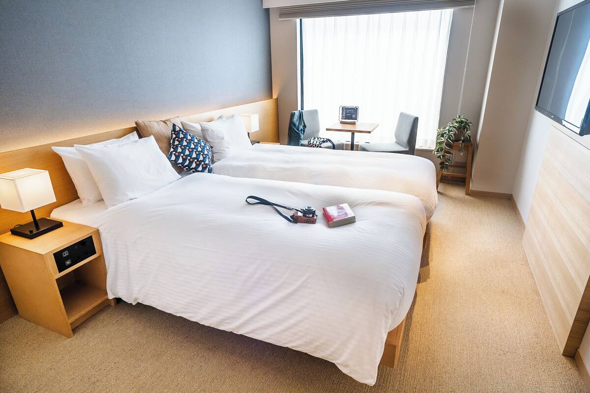 A hotel room with two beds, a bedside table with a lamp, a small table with a chair, and a window with curtains.