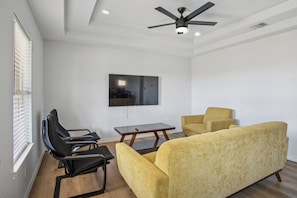 A modern living room with a ceiling fan, a flat-screen TV mounted on the wall, and a seating area with two armchairs and a sofa.