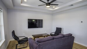A room with a ceiling fan, a flat-screen TV mounted on the wall, a purple sofa, two black chairs, and a wooden coffee table.