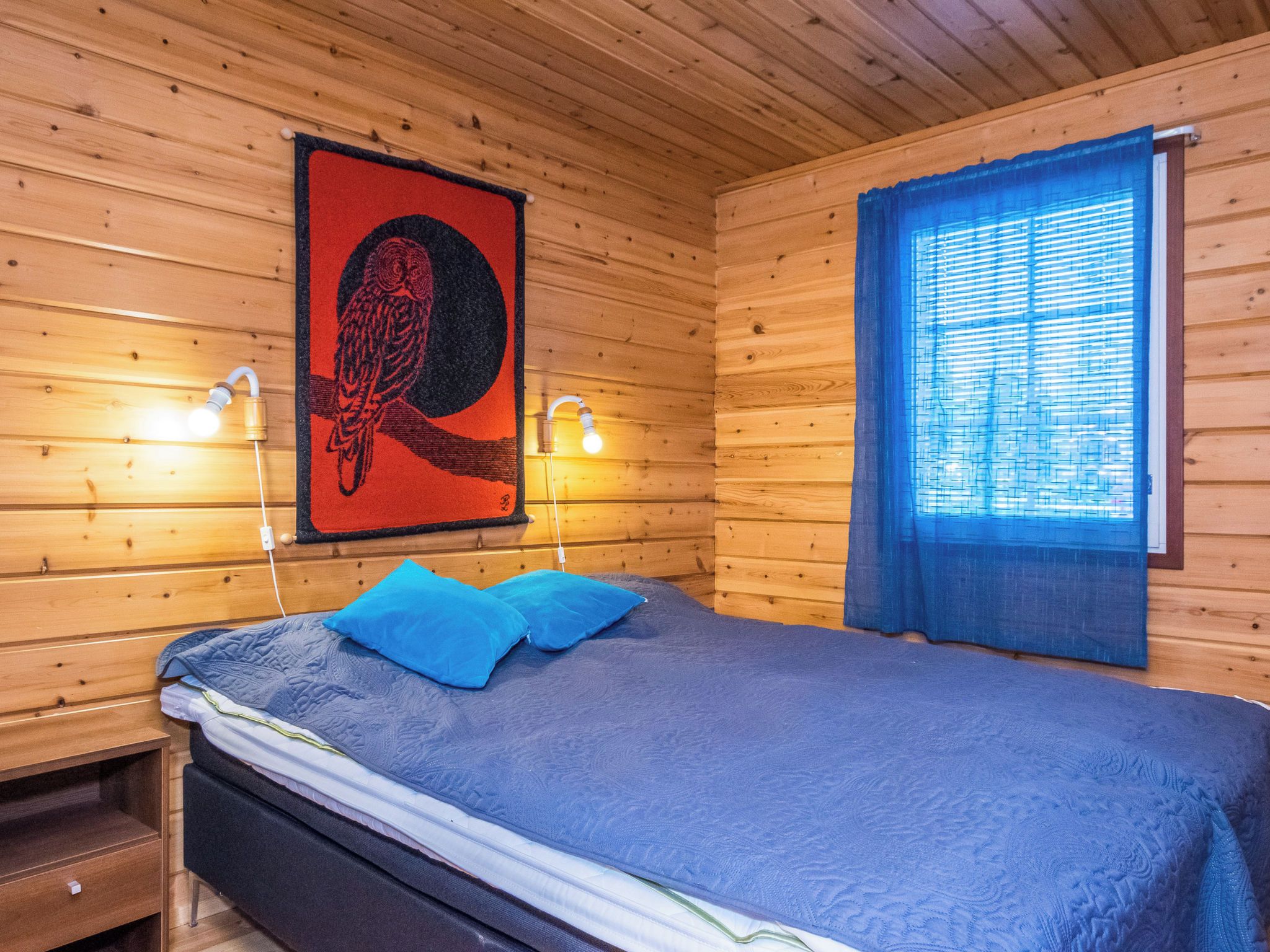 Een houten kamer met een bed, twee wandlampen, een raam met zonwering en een wanddecoratie.