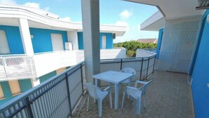 Une terrasse extérieure couverte, avec une table et des chaises, une rambarde en métal et une vue sur un bâtiment aux murs bleus et blancs.