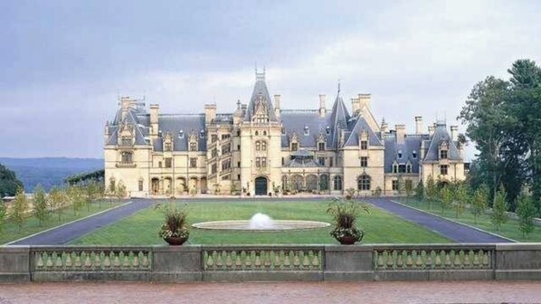 A large, historic building with a symmetrical facade, a central entrance, and a formal garden with a fountain.