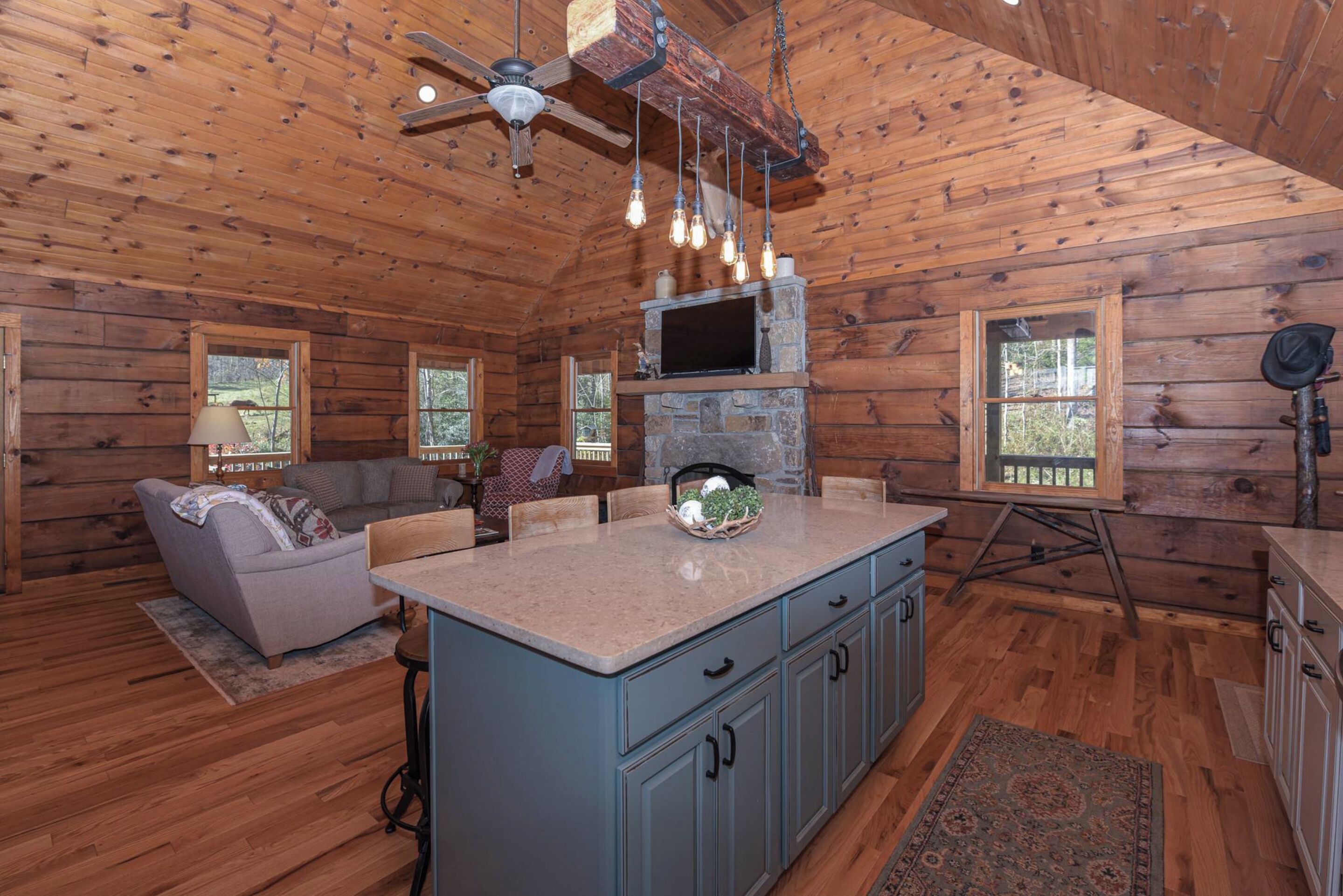 A cabin interior with a stone fireplace, wooden walls, a blue kitchen island, and a ceiling fan.
